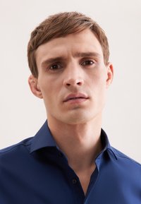 Jonge man met kort bruin haar in een donkerblauw overhemd met kraag, die recht vooruit kijkt met een neutrale uitdrukking op een lichte achtergrond.