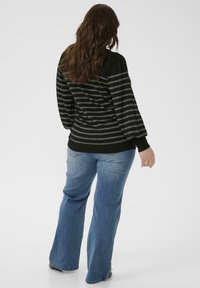 Mujer con cabello largo castaño, vistiendo un suéter a rayas negro y gris y vaqueros acampanados azules, de pie contra un fondo blanco liso.