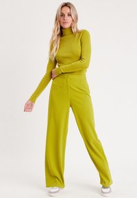 Un pull à col roulé vert vif et un pantalon large en tissu doux. Le pantalon a un bouton à l'avant et des poches. Des baskets avec des accents pastel.