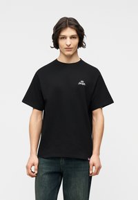 Νεαρό άτομο με μεσαίου μήκους σκούρα μαλλιά φοράει μαύρο κοντομάνικο μπλουζάκι με λευκό λογότυπο στο στήθος και μπλε τζιν, στέκεται μπροστά σε λευκό φόντο.