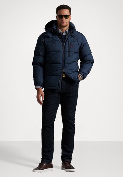 Mand iført en mørkeblå polstret jakke, stribet skjorte, mørke jeans, brune lædersneakers og solbriller, stående med den ene hånd i lommen.