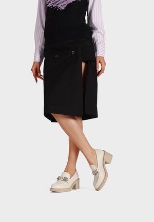 Zwarte asymmetrische rok met knoopdetail, gecombineerd met beige loafers met chunky hakken en een decoratieve metalen ketting. Geweven stof.