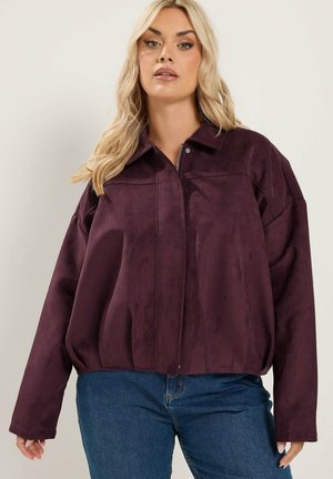 Frau trägt eine lockere dunkelviolette Reißverschlussjacke mit Kragen und blaue Jeans, steht vor einem schlichten hellen Hintergrund.