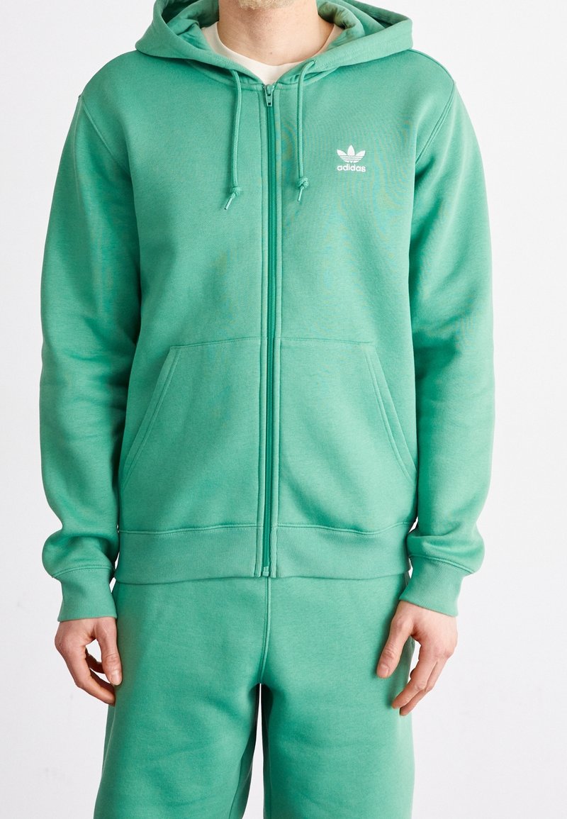 Man som bär en grön Adidas-hoodie och matchande svettbyxor, stående mot en helt vit bakgrund med händerna avslappnat vid sidorna.