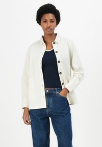 Off-white knoopjack met een kraag, twee voorzakken en bruine knopen, gecombineerd met een marineblauw tanktop en donkerblauwe jeans.