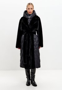 Zwarte lange jas met nepbont kraag, geborduurd patroon en een riem. Beschikt over een grijze capuchon als binnenlaag en slanke zwarte kniehoge laarzen.