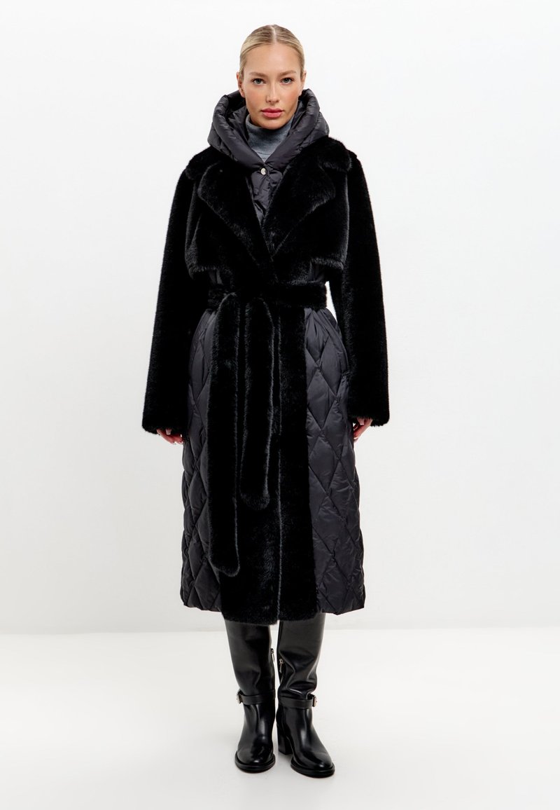 Zwarte lange jas met nepbont kraag, geborduurd patroon en een riem. Beschikt over een grijze capuchon als binnenlaag en slanke zwarte kniehoge laarzen.