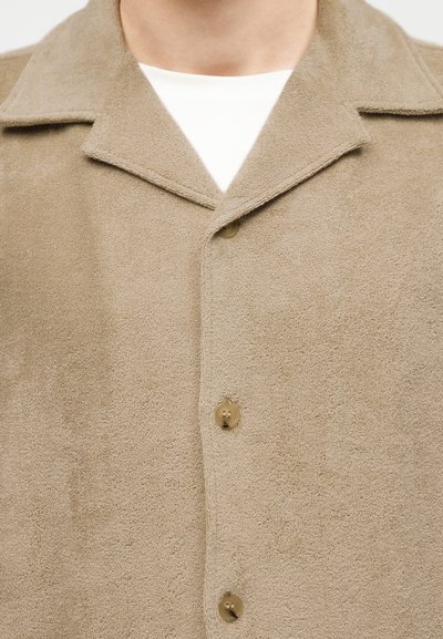 Κοντινό πλάνο πουκάμισου με υφή σε μπεζ χρώμα, με φαρδύ γιακά, φορεμένο πάνω από λευκό τοπ με στρογγυλή λαιμόκοψη.