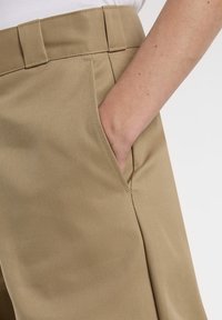 Kaki byxor med en slät, texturerad tyg och rak passform. En hand är placerad i sidofickan. Skräddarsydd midjebanddetalj.
