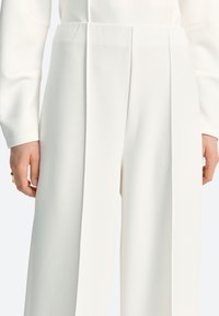 Balti plisēti bikses no gludas auduma ar augstu vidukli un platām kājām. Dizainā iekļautas asās locījuma līnijas priekšpusē.