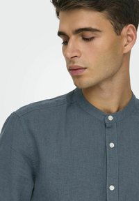 Camisa de lino gris con cuello Mao, cierre de botones y una textura sutil, situada sobre un fondo neutro.