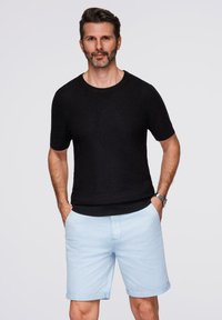 Maglione corto a maniche corte in tessuto nero abbinato a pantaloni corti azzurri. Il modello è in piedi con le mani nelle tasche, mostrando un abbigliamento casual.