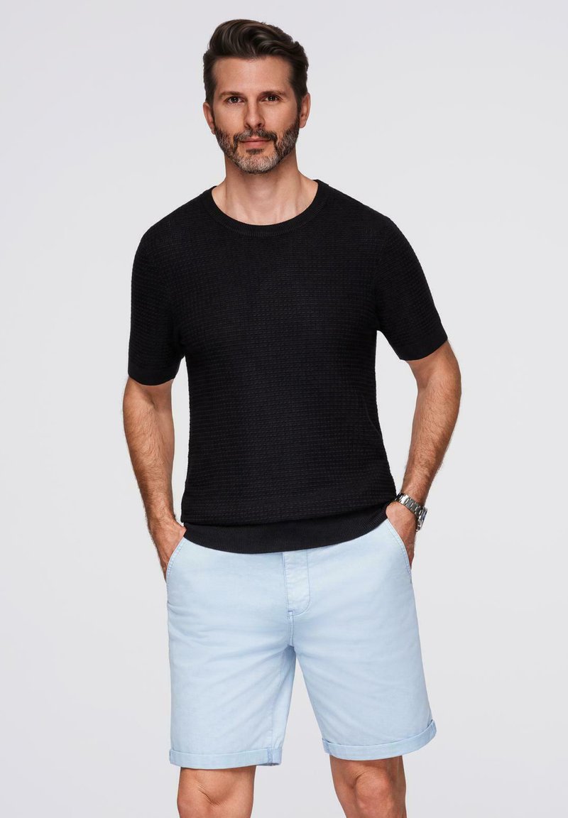 Maglione corto a maniche corte in tessuto nero abbinato a pantaloni corti azzurri. Il modello è in piedi con le mani nelle tasche, mostrando un abbigliamento casual.