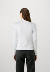 Hvit langermet topp med høy hals, med en bakre søm og et rosa logo-patch på venstre arm. Parret med mørke, rettskårne jeans.