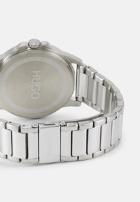 Montre en acier inoxydable argentée avec une finition lisse et polie, étanche jusqu'à 3 bars. Comprend un bracelet brossé et un logo gravé.