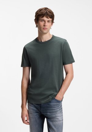 Jeune homme aux cheveux courts et bruns, portant un t-shirt vert foncé à manches courtes et un jean bleu, debout avec une main dans la poche.