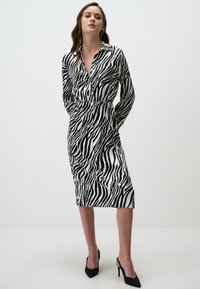 Zwart-wit gestreepte jurk met kraag, lange mouwen, elastische taille en middellange lengte. Draagt met zwarte hakken.