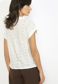 Balta īsroku blūze ar perforētu mežģīņu rakstu, apaļu izgriezumu un vilnainu apakšmalu. Audums izskatās viegls un teksturēts.