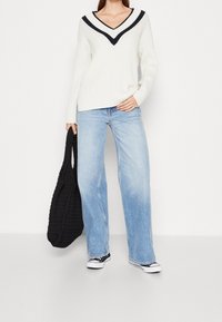 Vit ribbad tröja med svart V-ringning, kombinerad med ljusblå vida jeans. Modellen håller en svart vävd tote-väska och bär svarta sneakers.