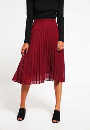 Femme portant un top noir à manches longues, une jupe midi plissée bordeaux et des mules à talons ouvertes noires, debout jambes écartées sur fond blanc.