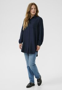 Chemise bleu marine à boutons avec manches longues, poche poitrine et coupe décontractée, assortie à un jean bleu clair et des chaussures noires.