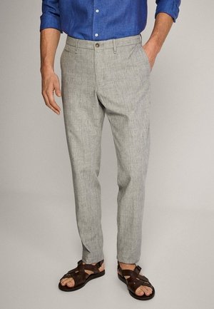 Uomo che indossa pantaloni di lino grigio chiaro, camicia blu con bottoni e sandali in pelle marrone scuro, in piedi con una mano in tasca.