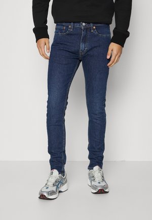 Hombre con vaqueros ajustados azul oscuro y zapatillas blancas y grises, de pie frente a un fondo gris claro.