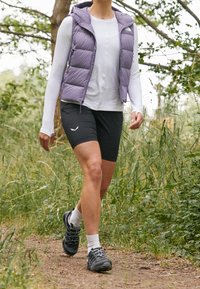 Pikowana lawendowa kamizelka noszona na białej koszuli z długim rękawem, w połączeniu z czarnymi szortami i ciemnymi sportowcami, spacerująca po ziemnej ścieżce otoczonej zielenią.