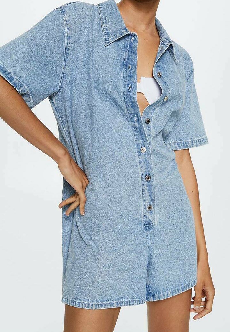 Femme portant une combishort en denim bleu clair à manches courtes avec des boutons sur le devant, une main sur la hanche, et un haut blanc visible en-dessous.