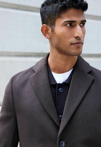 Mörkbrun kappa med skräddarsydd design, utrustad med notch-lapel och knäppning, lagd över en marinblå stickad skjorta med blå knappar.