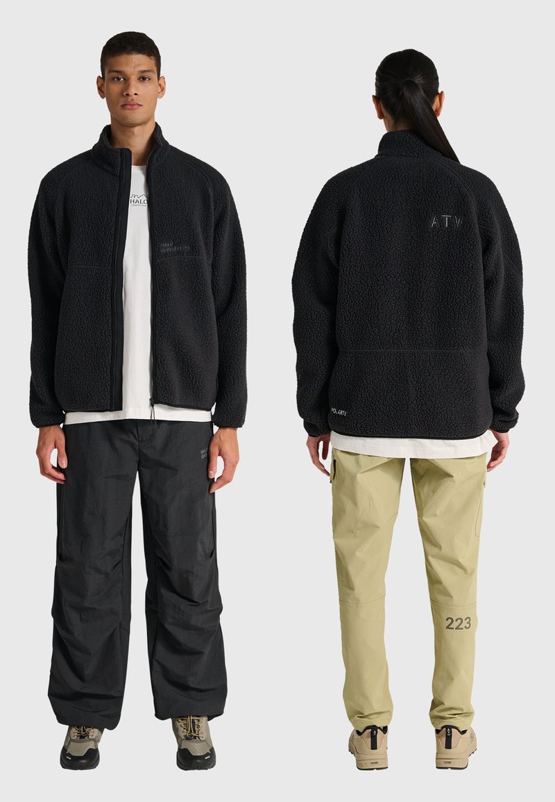 Zwei Models tragen schwarze Fleecejacken; eines zeigt die Vorderseite mit schwarzer Hose, das andere die Rückseite mit khakifarbener Hose und beigen Schuhen.