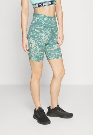 Kobieta ubrana w zielone, wzorzyste, wysokowciśnięte spodenki rowerowe Puma, czarne buty sportowe Puma oraz turkusowy sportowy biustonosz Puma.