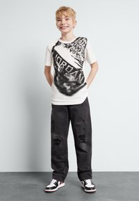 White t-shirt with black graphic pattern, paired with distressed black pants and black and white sneakers. Casual fit, short sleeves.