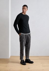 Suéter negro de punto con puños acanalados, combinado con pantalones de corduroy a rayas grises y zapatos negros de cordones sobre un suelo de madera.