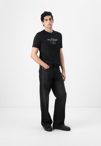 Zwarte katoenen t-shirt met witte logo-print, gecombineerd met brede donkere denim jeans. Model staat voor een neutrale achtergrond en draagt zwarte schoenen.