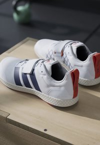 Biele športové topánky s námorníckymi pruhmi, textúrovanou látkou a červeným akcentom na päte. Obsahujú suchý zips a vzorovanú gumenú podrážku.