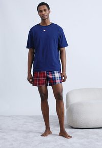 Μπλε βαμβακερό μπλουζάκι με μικρό λογότυπο, συνδυασμένο με βαμβακερά καρό σορτς σε κόκκινο, μπλε και λευκό. Το μοντέλο στέκεται σε ανοιχτόχρωμο χαλί κοντά σε έναν απαλό, καμπυλωτό καναπέ.