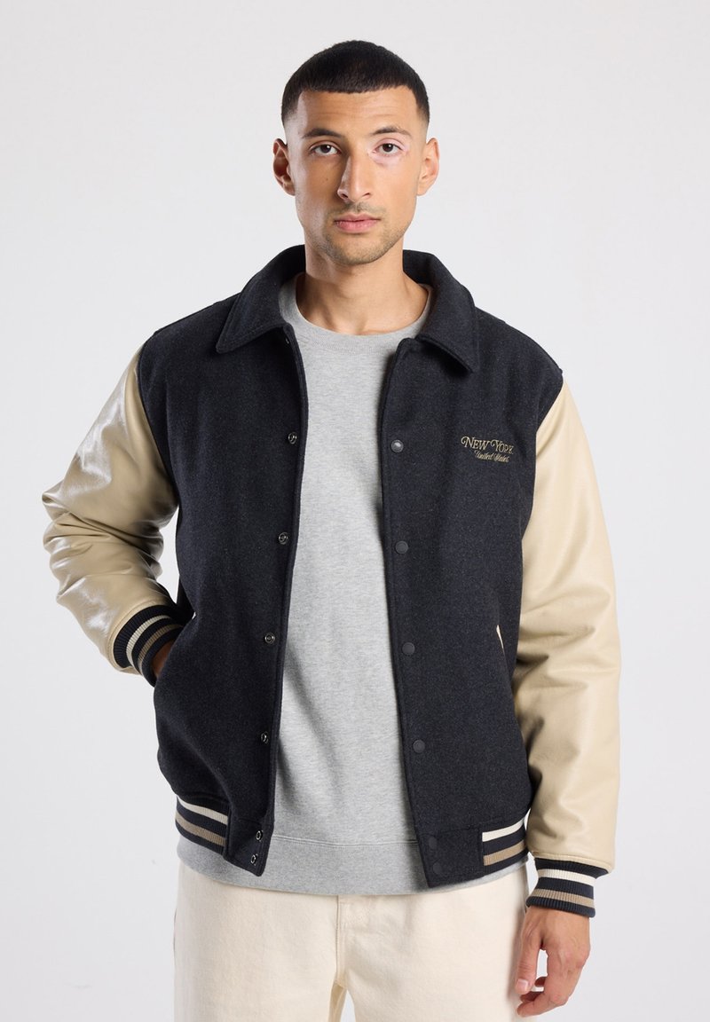 Wool-blend varsity jacket with black body, beige faux-leather sleeves, ribbed striped cuffs, and embroidered "NEW YORK" detail on chest.