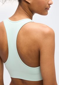 Γυναίκα που φοράει ανοιχτό πράσινο αθλητικό σουτιέν με σχεδιασμό racerback, δείχνοντας τον ώμο και το πάνω μέρος της πλάτης της ενάντια σε ένα απλό φόντο.