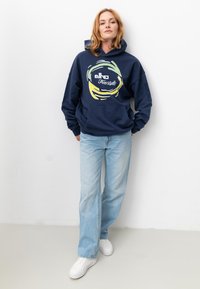 Dunkelblauer Kapuzenpullover mit einer Vordertasche, der ein kreisförmiges Grafikdesign in Gelb und Grün zeigt. Kombiniert mit hellblauen Jeans und weißen Turnschuhen.