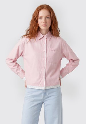 Junge Frau mit roten Haaren, die ein hellrosa Hemd mit Knöpfen und hellblaue Jeans trägt, steht mit den Händen in der Hüfte vor weißem Hintergrund.
