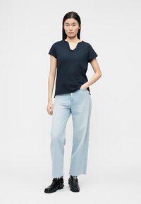 Νεαρή γυναίκα στέκεται με το ένα χέρι στην τσέπη, φοράει μαύρο κοντομάνικο πουκάμισο, ανοιχτόχρωμα φαρδιά τζιν παντελόνια και μαύρα μποτάκια στον αστράγαλο.