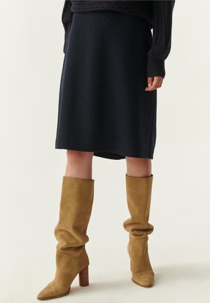 Jupe en tricot bleu marine avec un design droit, associée à des bottes en daim beige clair, hautes jusqu'au genou, avec un talon block et un bout pointu.
