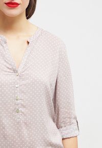 Blusa grigia chiaro con scollo a V, chiusura a bottoni e maniche arrotolate. Presenta un delicato motivo floreale bianco su un tessuto liscio.