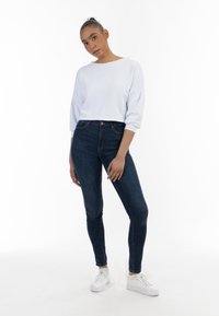 Felpa bianca a maniche a pipistrello, abbinata a jeans skinny blu scuro e sneakers bianche su uno sfondo neutro.
