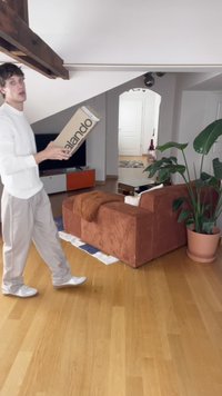Scatola di cartone con il logo "Zalando", tenuta da una persona in un maglione bianco; presenta un divano in velluto a costine marrone, una pianta verde e un pavimento in legno.