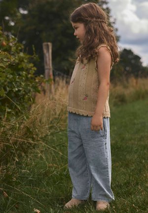Ung jente med krøllete hår står barfot på gresset, iført en strikket beige vest og løse blå jeans, ser på planter nær et gjerde av tre.