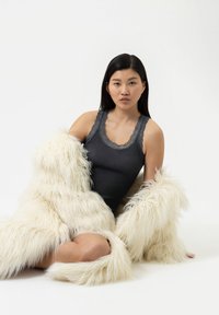 Grijze geribde tanktop met kantafwerking, gecombineerd met een lange witte nepbontjas met een zachte, fluffy textuur. Model zit met gekruiste benen.