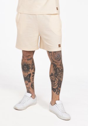 Man draagt crèmekleurige korte broek en witte sneakers, met getatoeëerde benen met bloemen- en zonontwerpen, tegen een effen witte achtergrond.