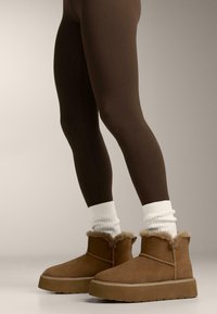 Brune ruskinds-ankelstøvler med fluffet, tan pelskant og tyk gummisål, båret med ribstrikkede hvide sokker og brune leggings.